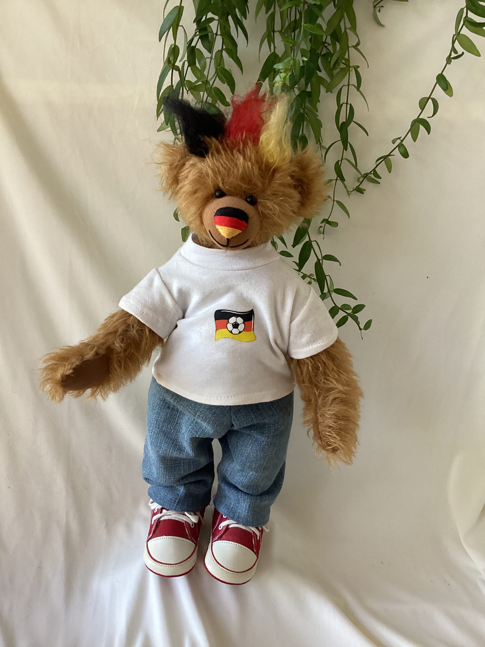 contact-img Ein handgenähter Teddybär steht stolz vor einer weißen Stoffkulisse mit grüner Hängepflanze. Er trägt ein Fußball-Fan-Outfit mit Deutschlandflagge, Jeans und roten Sneakers. Die bunten Fellsträhnen in Schwarz-Rot-Gold und das fröhliche Gesicht verleihen ihm einen charmanten, mitreißenden Charakter.