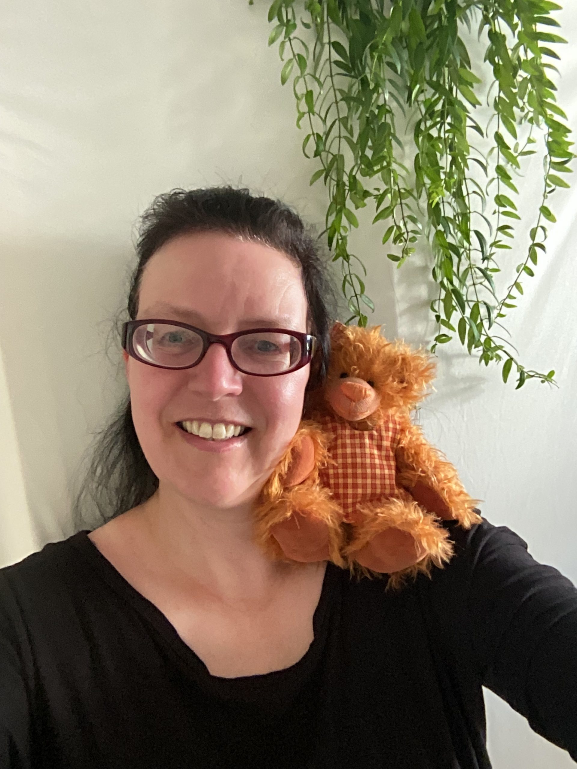 our-services-1 Eine lächelnde Frau mit dunklem Haar und Brille trägt liebevoll ein handgenähtes, orangefarbenes Teddybären Mädchen mit kariertem Latz auf der Schulter. Die beiden strahlen vor einer weißen Wand mit grüner Hängepflanze eine warme, kreative Atmosphäre aus.